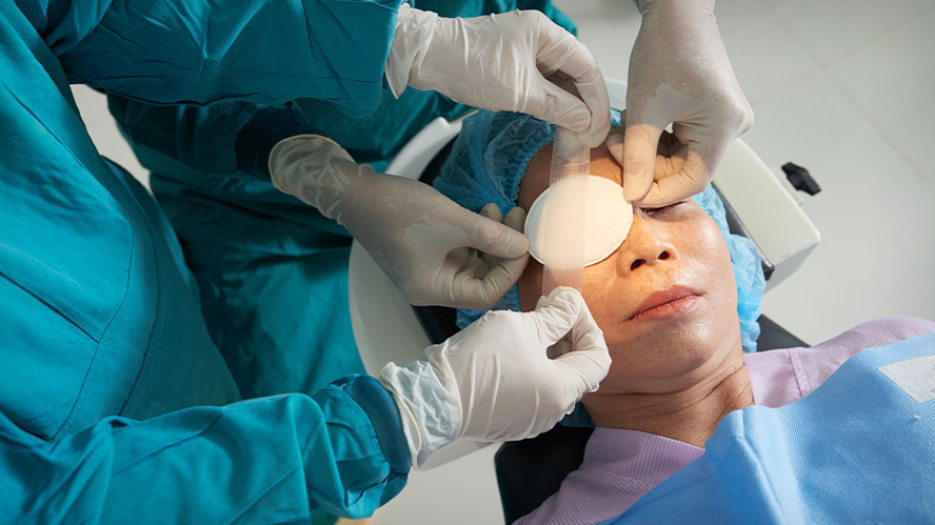 Two surgeons, in teal gowns with white gloves, placing eye patch on reclined older female patient.